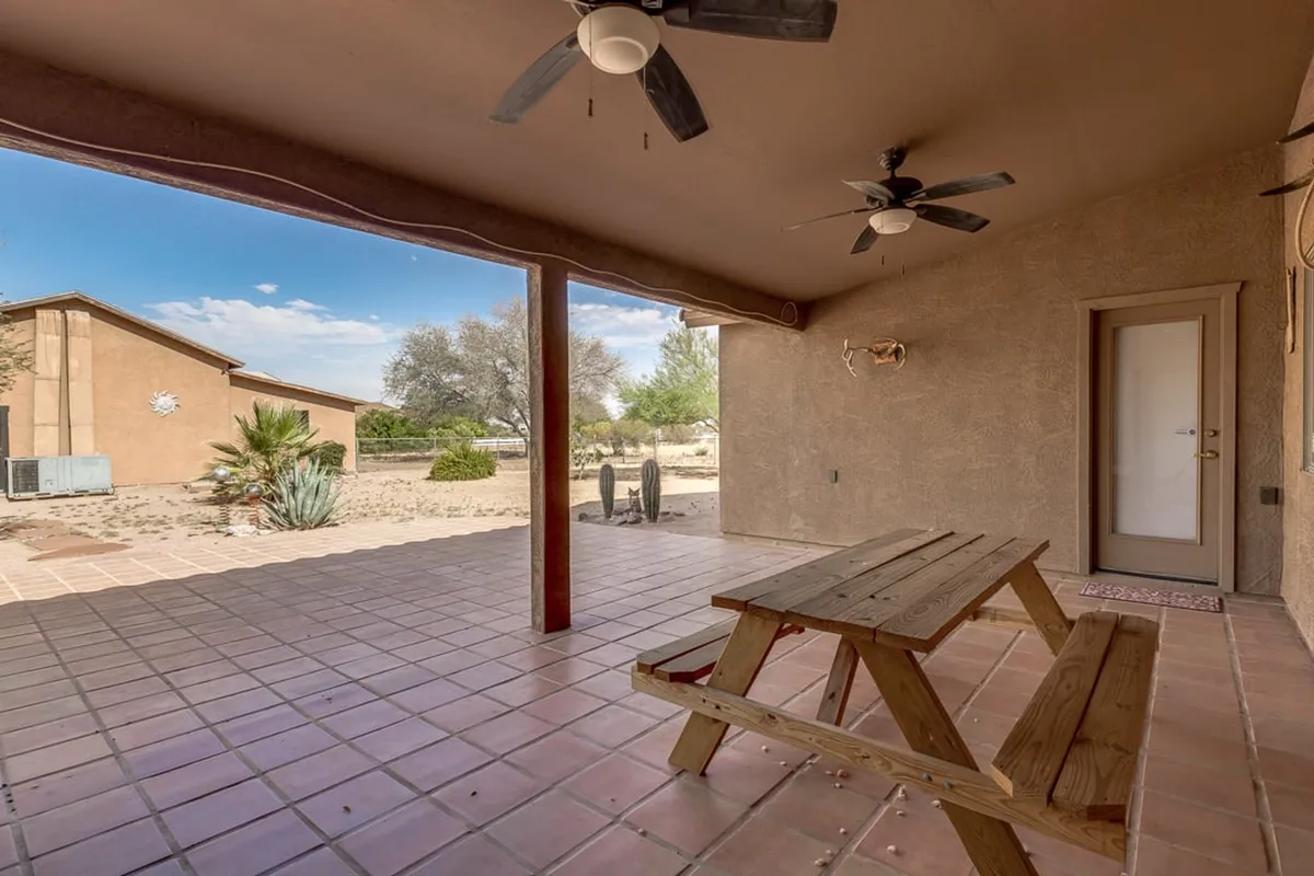 A spacious covered patio with a picnic table and ceiling fan overlooks a scenic landscape. The area invites outdoor dining, group relaxation, and family gatherings under the open Arizona sky.