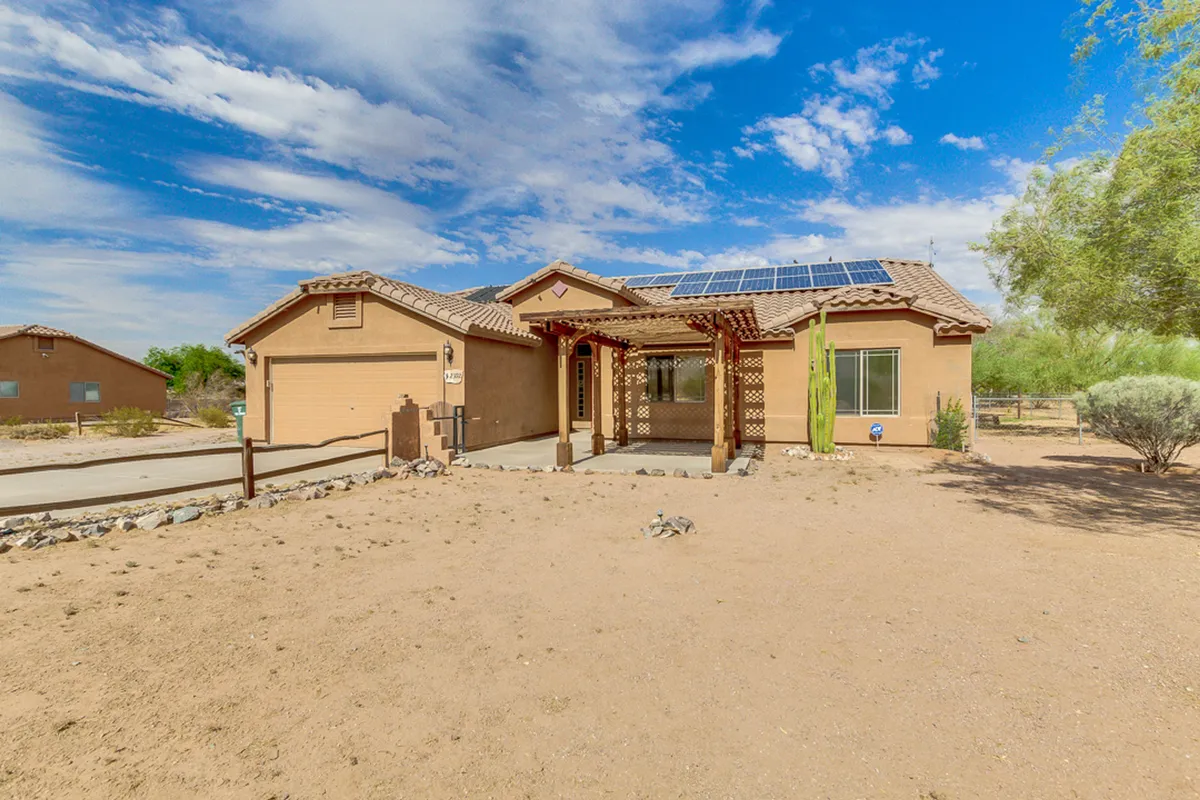 This image showcases a welcoming front porch with solar panels visible on the roof, surrounded by a classic Arizona desert landscape complete with cactus. The setting sun casts a warm glow, and the porch suggests an inviting space to relax and enjoy the quiet, open surroundings.