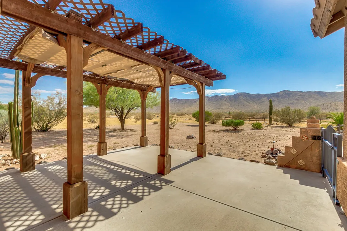 A wooden pergola patio sits at the edge of a desert landscape, with lush green vegetation framing open views of distant mountain silhouettes. The setting sun casts a golden light over the scene, promising peaceful moments spent relaxing, socializing, or simply taking in the scenery.
