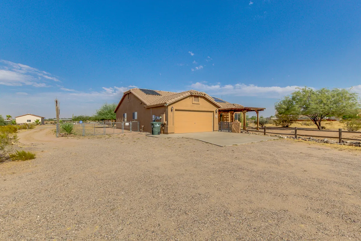 The house’s facade, garage, and front porch are surrounded by a natural desert landscape. The view extends to the horizon, with the terrain suggesting the proximity of hiking or riding paths. The inviting porch seamlessly bridges indoor comfort and outdoor exploration.