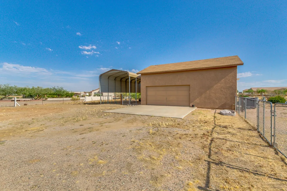 An exterior shot showcasing a garage structure, adjacent carport, and open land. The wide-open yard and relaxed ambiance emphasize the property's spaciousness and potential for accommodating horse trailers and easy access to trails.