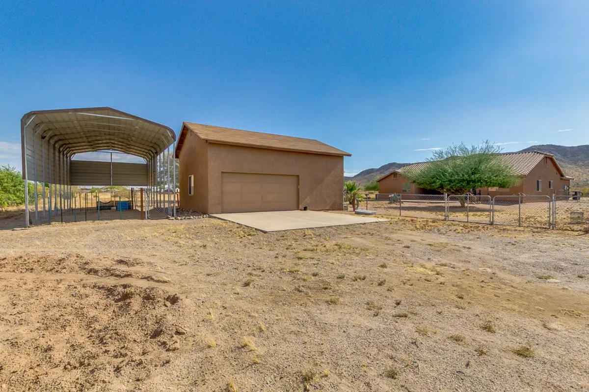 The main house is pictured with a carport and large fenced area, set against a dramatic mountain backdrop. The expansive outdoor space suggests ample room for parking RVs, horse trailers, or ATVs, with wide-open prairie extending beyond.