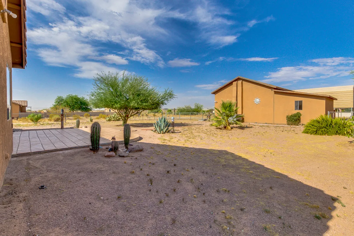A tranquil scene of the house exterior sitting amidst desert flora, including cacti and drought-resistant plants. The laid-back mood and open yard suggest space for outdoor gatherings or launching outdoor excursions, with sunset colors subtly painting the sky.